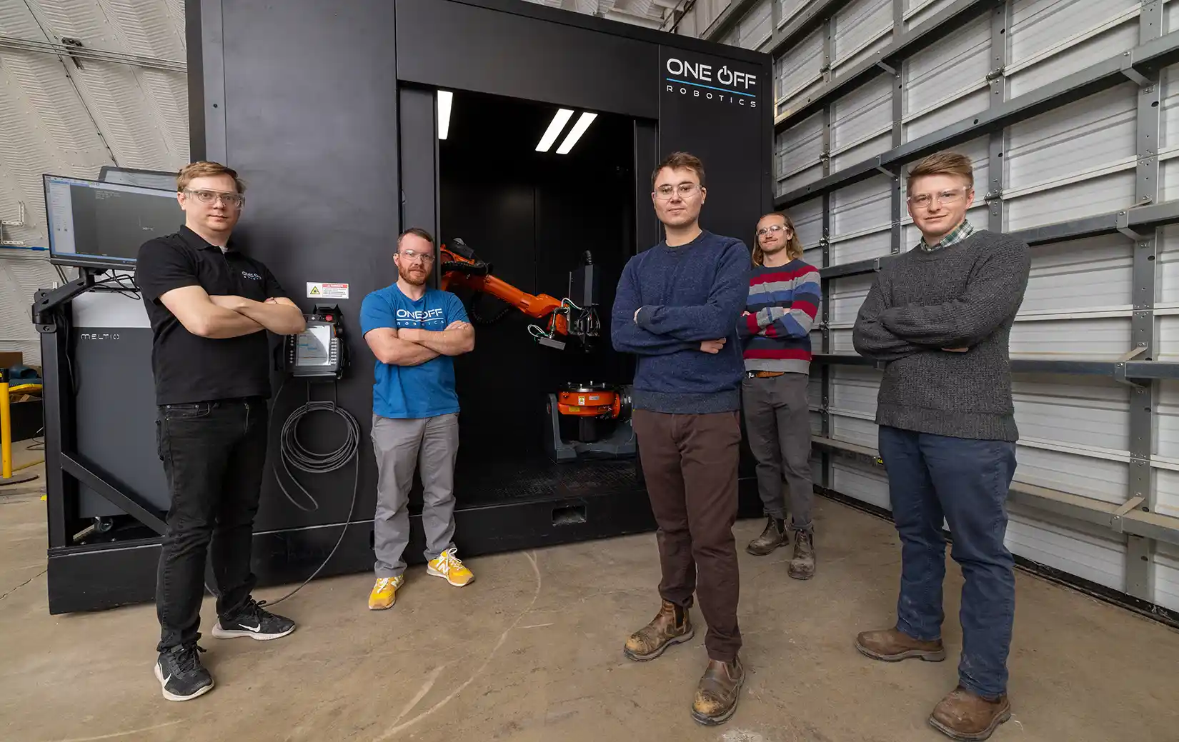 Five people stand outside a big black box housing an orange robot arm and table.
