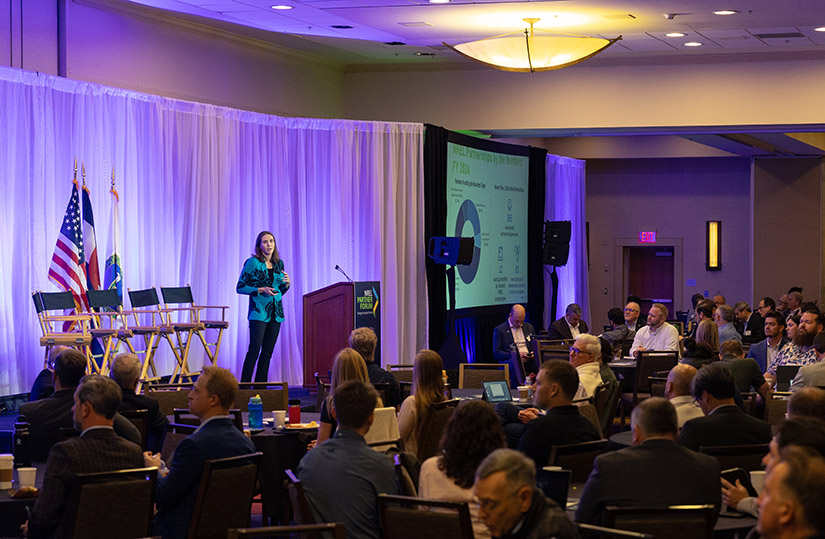 A person on a stage speaks to a room of people.