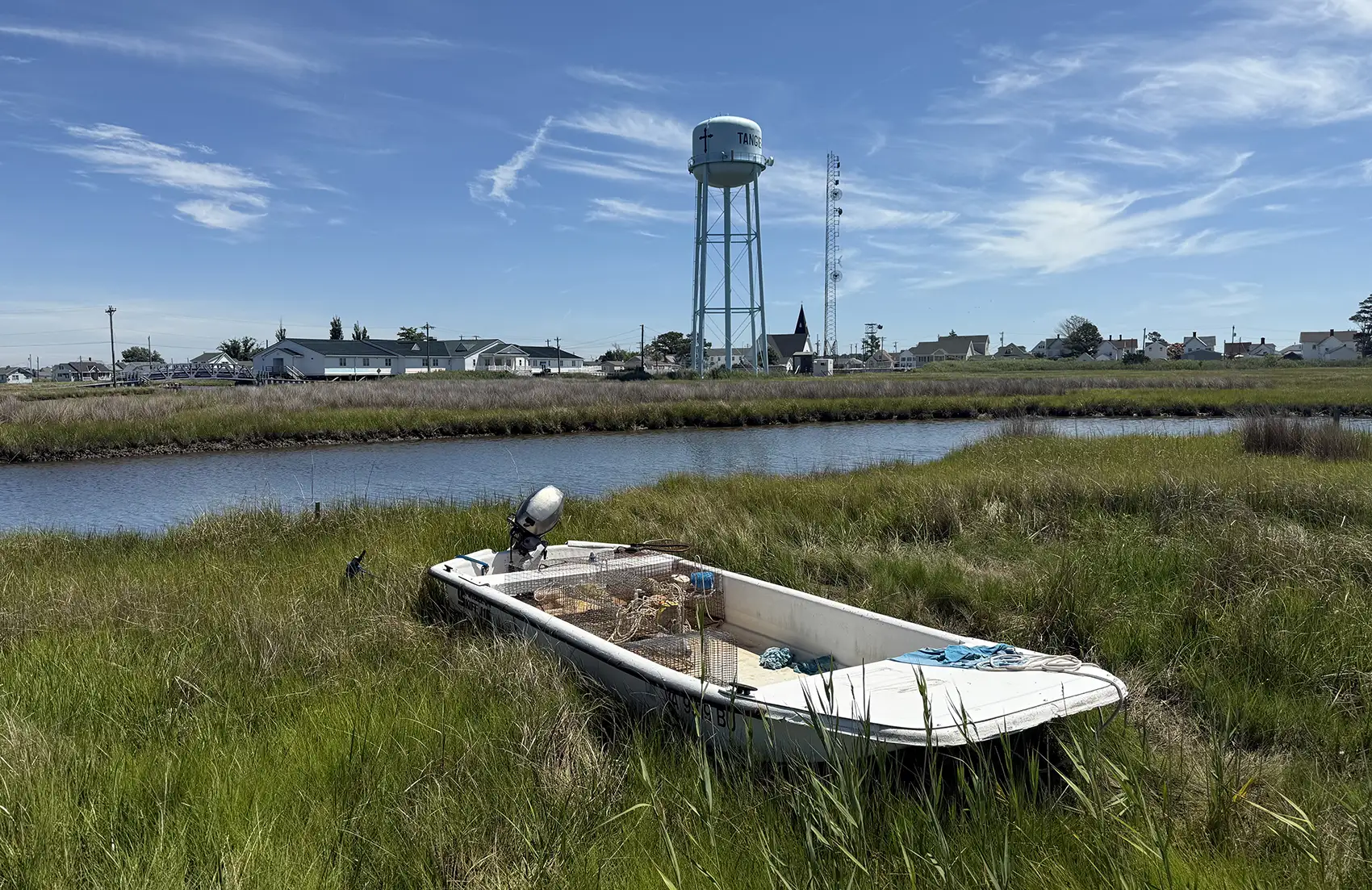 A small motor boat with crabbing equipment sits on a grassy area in front of a waterway with a water tower behind it.