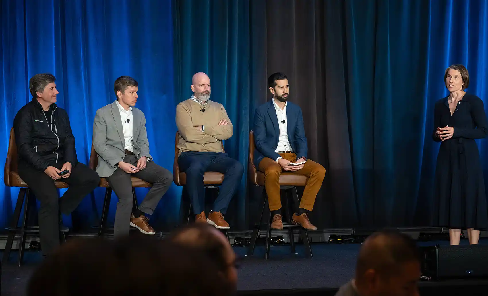 Four men in business attire sit side by side onstage during a panel discussion, set against a blue-lit curtain backdrop.