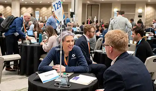 Continue reading about CEO and investor sit across from each other at a small networking table, engaged in conversation, surrounded by a large, crowded room filled with hundreds of attendees having similar one-on-one discussions at mini tables.
