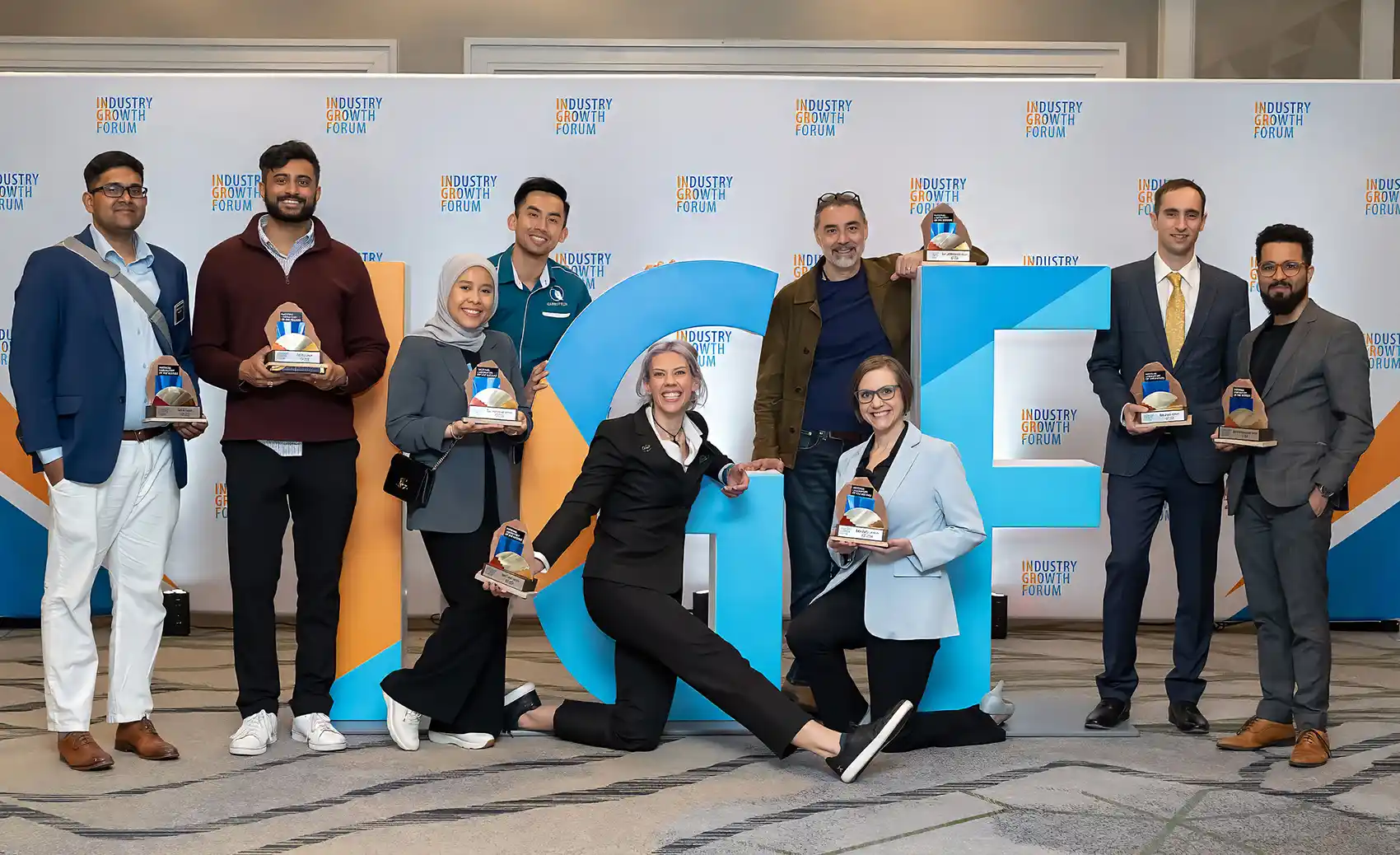 Nine people stand in front of an IGF step-and-repeat banner, arranged around large five-foot-tall “IGF” letters, holding awards in celebration.