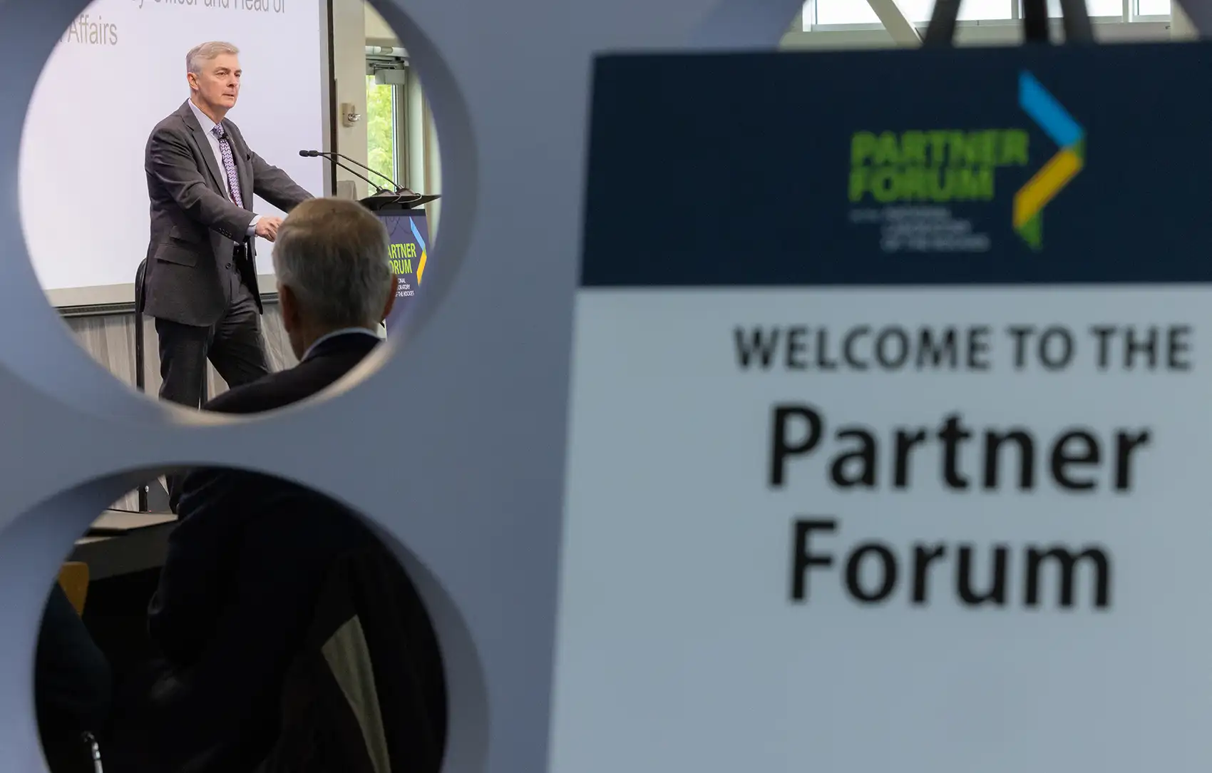 A person speaking at a podium with a sign with the text Welcome to the Partner Forum in the foreground.
