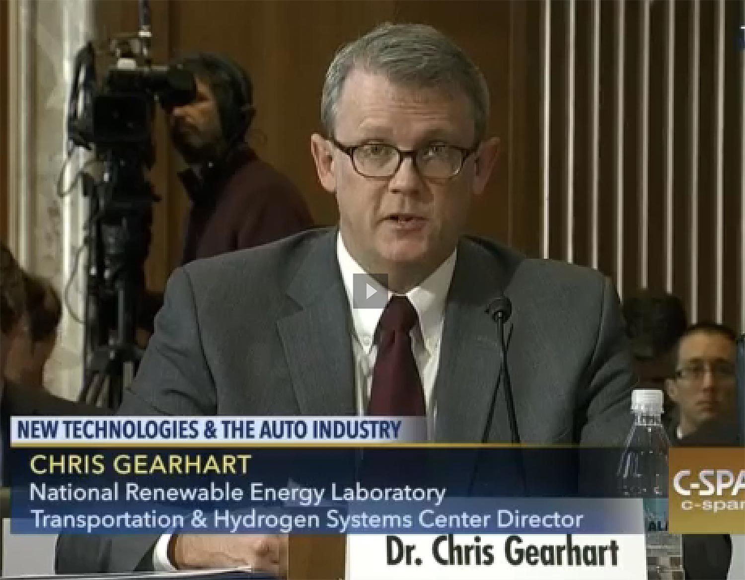 Photo of a man wearing glasses and in a suit sitting in front of a nameplate that says: