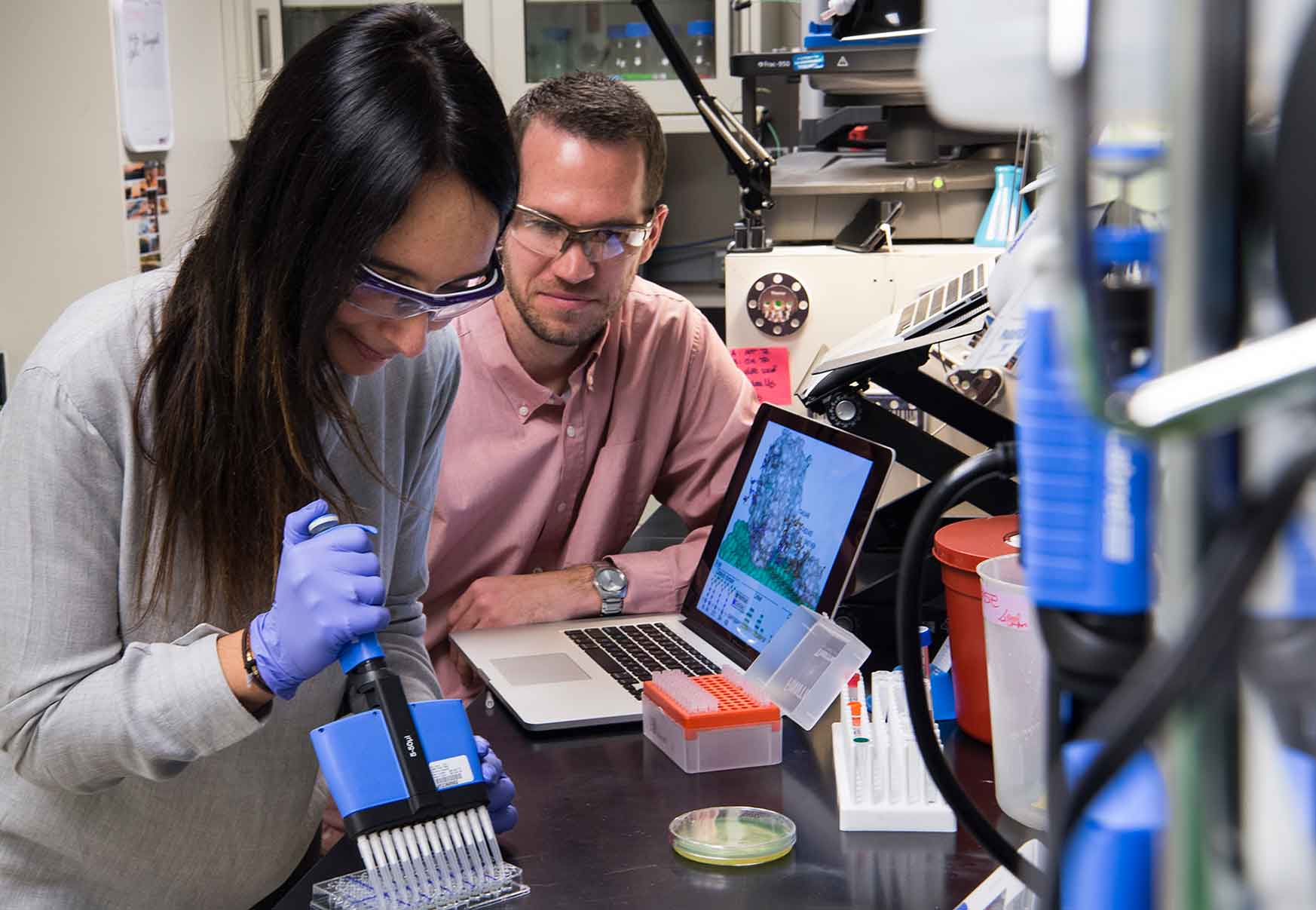 One woman and one man work at a laboratory bench surrounded by lab equipment.