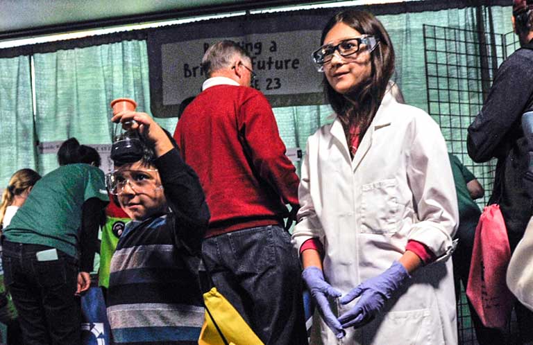 Photo of a young girl wearing a large lab coat. A small boy stands next to her holding a beaker of green liquid.