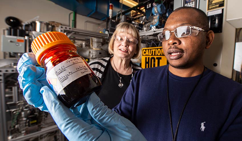 One scientist in a laboratory holds a bottle of a dark liquid in his hands while another scientist looks over his shoulder.