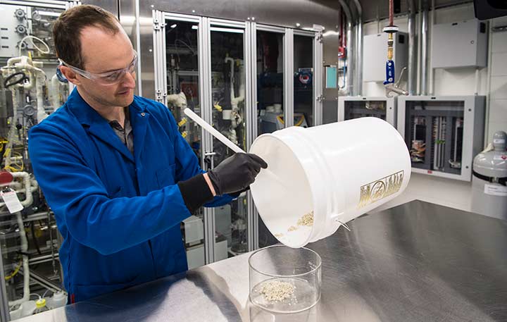 A man in a lab coat pours catalyst pellets from a bucket into a plastic beaker.