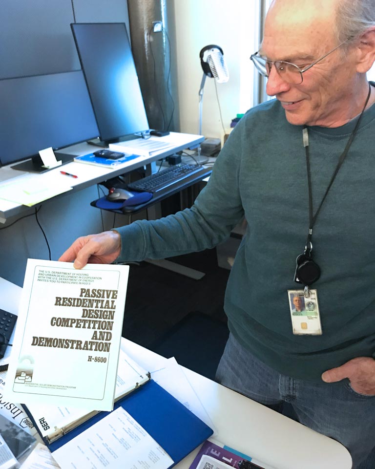 Ron Judkoff stands in an office holding a copy of a manual titled 