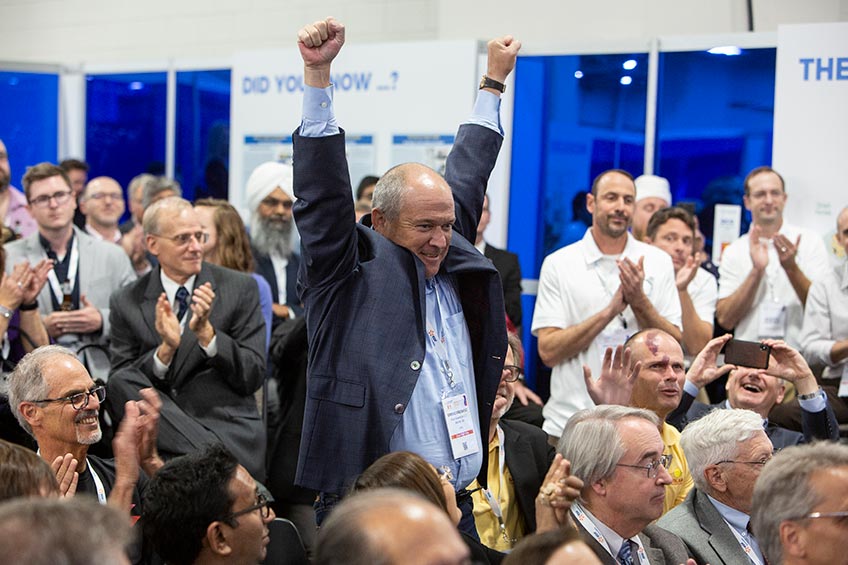 A man stands with his arms up in a crowd of people sitting down after winning a contest.
