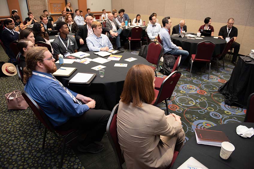 A group of people sit at tables listening to a presenter.