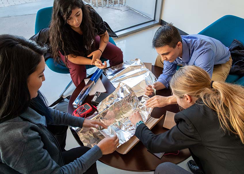 Three women and a man work on a piece of electronics at a table.