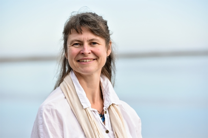 A woman, Genevra Harker-Klimeš, smiles at camera in front of a blurred blue background.