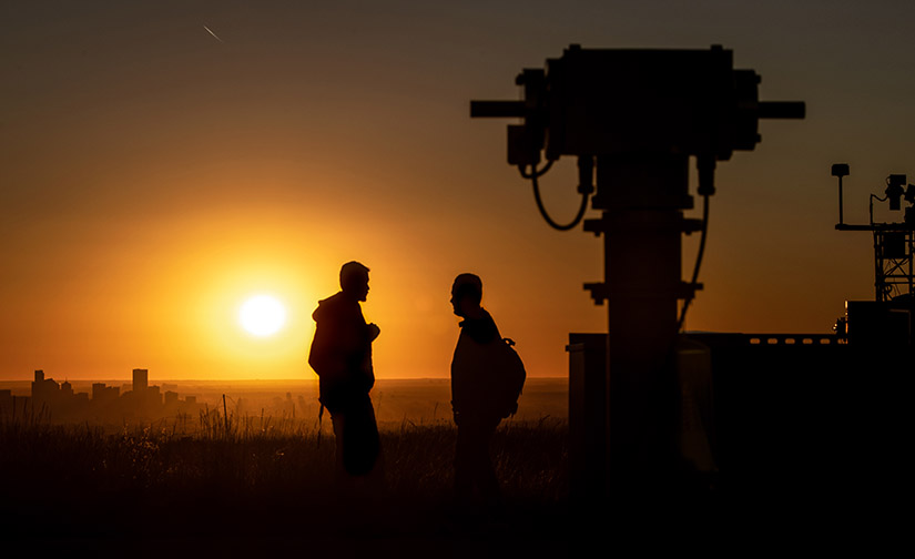 Two men speak outdoors with the Denver skyline and sunrise in the background