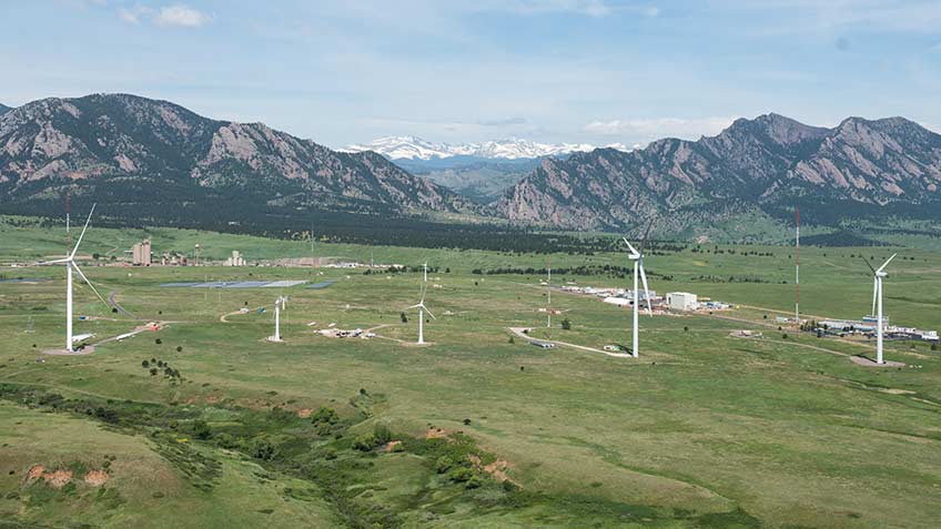 An aerial photograph of a energy laboratory campus with a wind turbine in the foreground and mountains in the background on a clear day. 