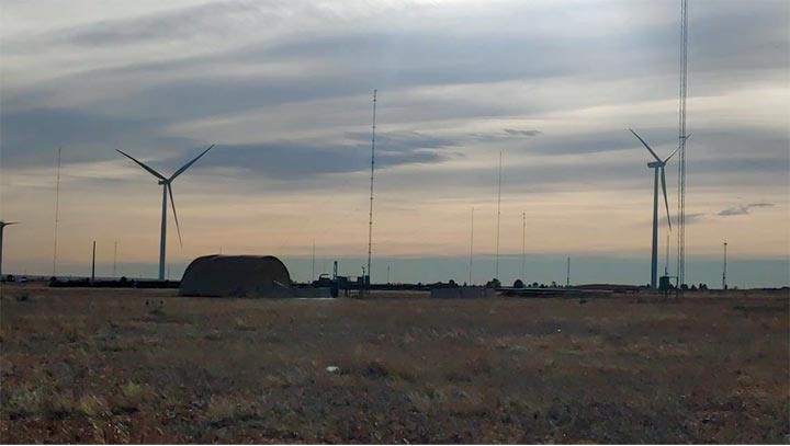 An open landscape at twilight with wind turbines and a Quonset hut in the distance