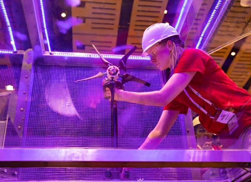 A student in a hard hat and safety glasses adjusts a model wind turbine inside a wind test tunnel.