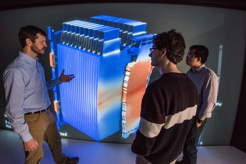 Three researchers stand in front of a large computer screen with the image of a battery.