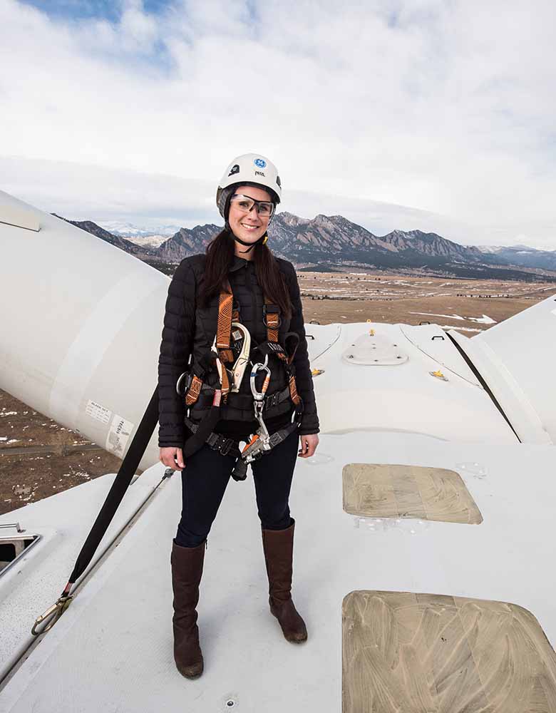 A woman standing on top of a wind turbine in goggles, hard hat, and harness.
