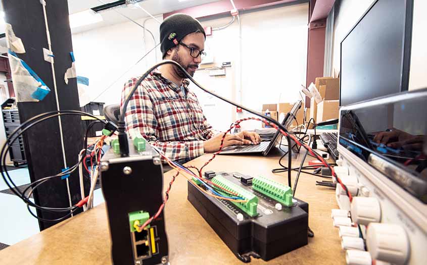 A man working on a laptop next to electronics and wiring.