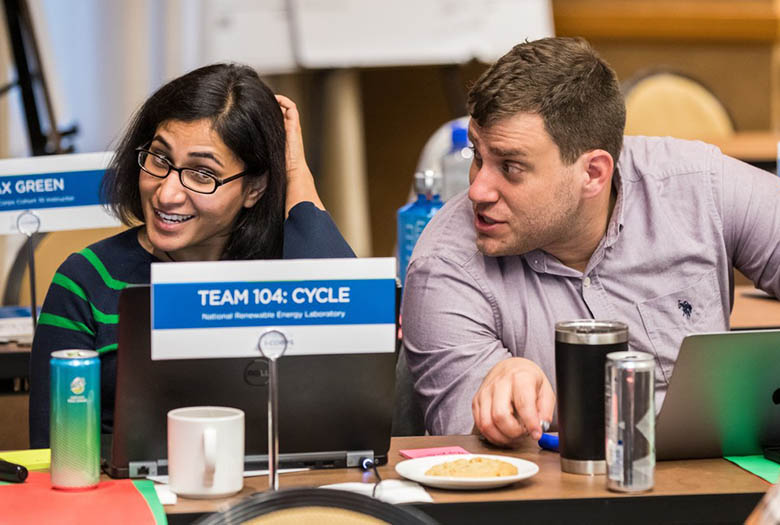 Avantika Singh and Nic Rorrer sit and chat at a table behind a sign that reads: 