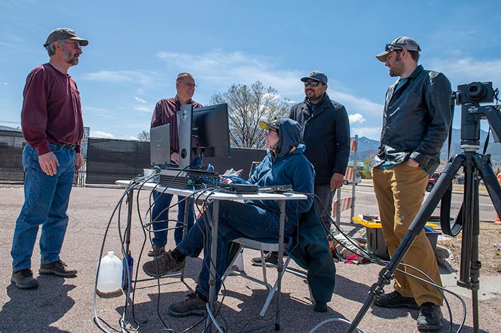 Photo of four people chatting in outside setting near a person sitting at a table in front of a computer.
