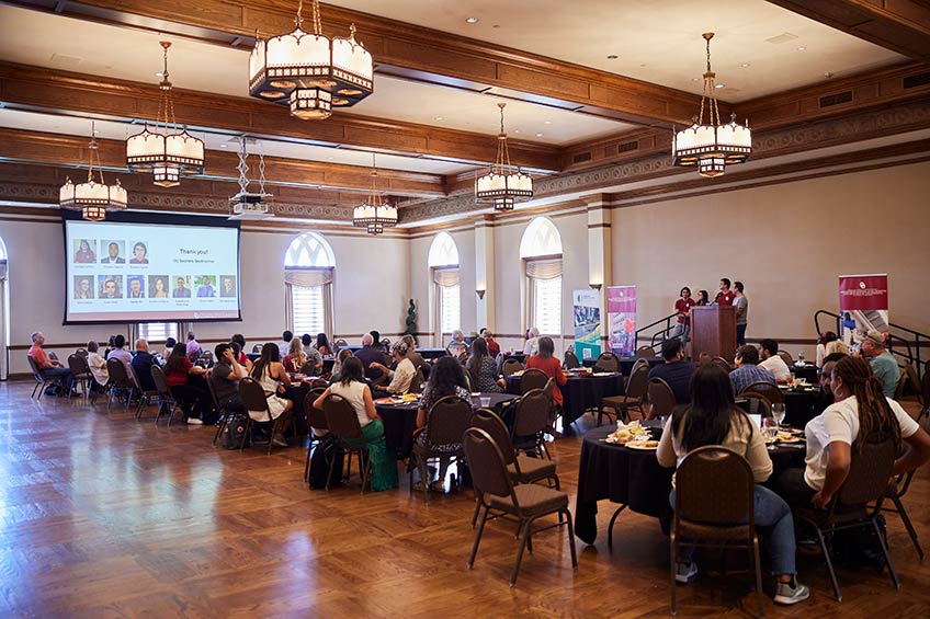 Dozens of people fill round tables in a ballroom and listen to a presentation from four students.