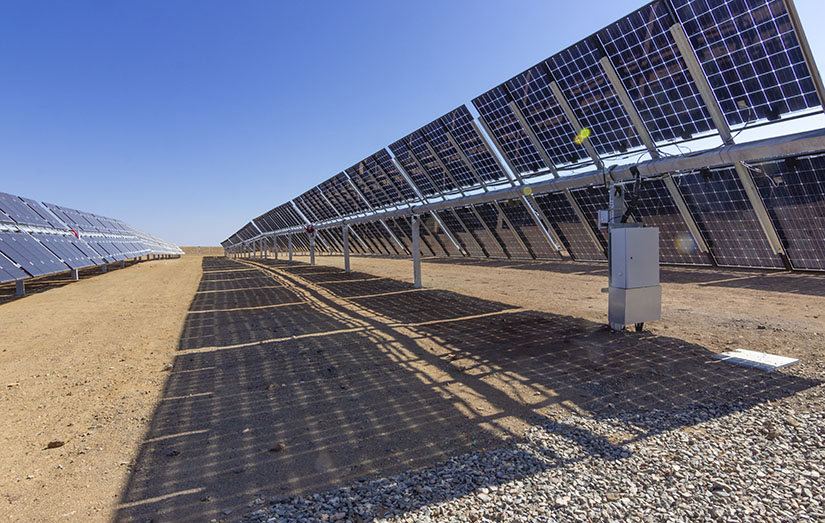 A row of solar panels casts a shadow on a dirt field on a sunny day.