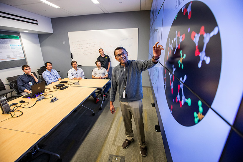 A researcher stands in front of a large screen. Five other researchers sit in the background. 