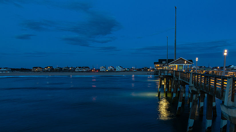 The ocean during sunrise with the beach and houses in the background, a pier on the right side