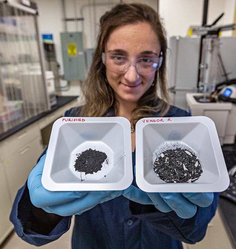 A scientist in lab coat, safety goggles, and gloves holds two sample containers of debris, the one on the right a mix of sizes and colors