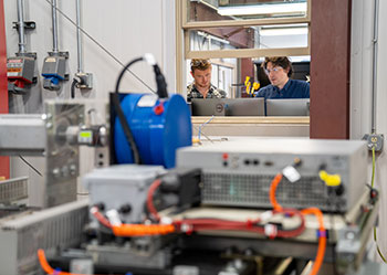 Two people look at computer screens with a stack of metal hardware and wires in the foreground