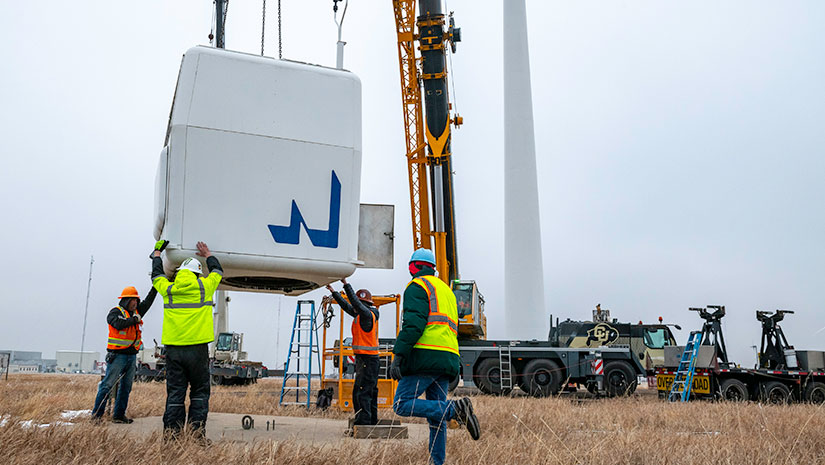 Crews wearing protective gear position a large wind turbine component being lowered by a crane.
