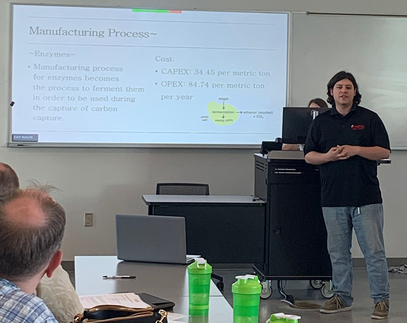 A student stands at the front of a classroom presenting with a slide on a screen behind him.