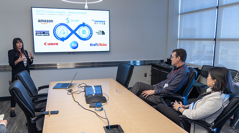 Two people in a conference room facing a presenter standing in front of a presentation screen.