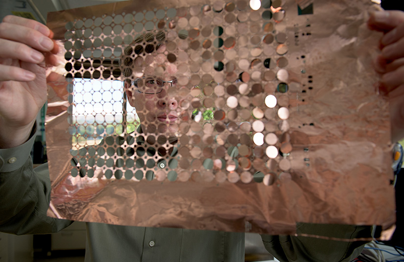 A man holds a sheet of copper discs in front of his face. Some of the sheet has empty holes where the discs have already come out.