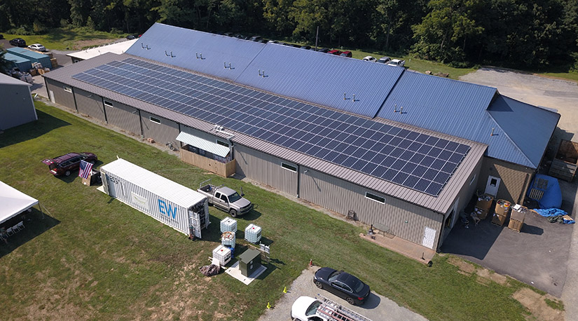 A white storage container sits on grass next to a larger building with solar panels on top.