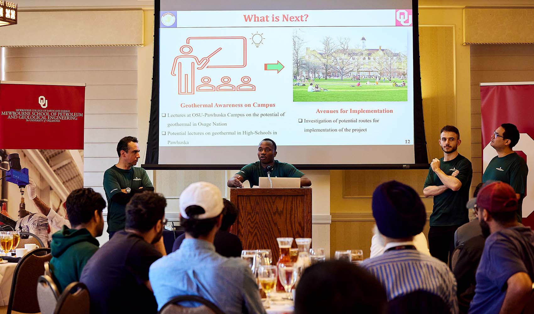 One person speaks at the podium to a crowd with three other team members beside them.