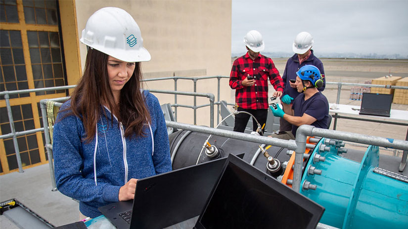 A person in a hard hat stands at a laptop in front of a big piece of hydropower machinery; three people in hard hats review something together