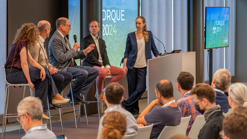 Four people sit and one person stands at a podium on a stage with screens and audience members visible in the foreground