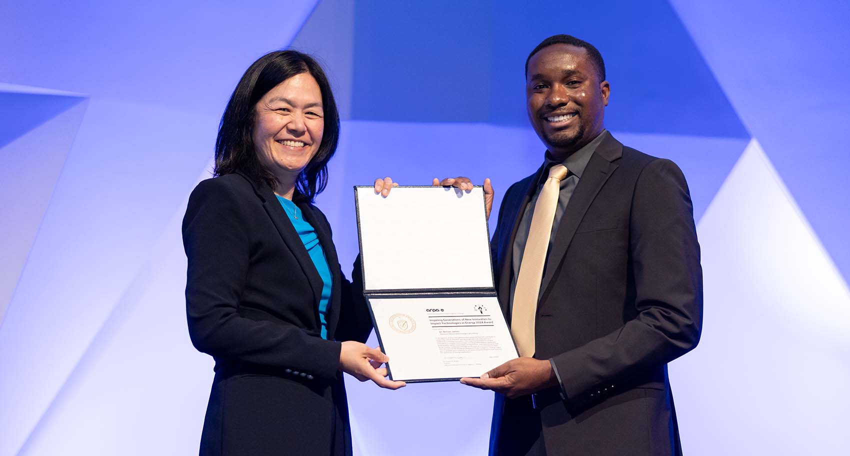 two people stand on a stage holding a certificate and smiling 