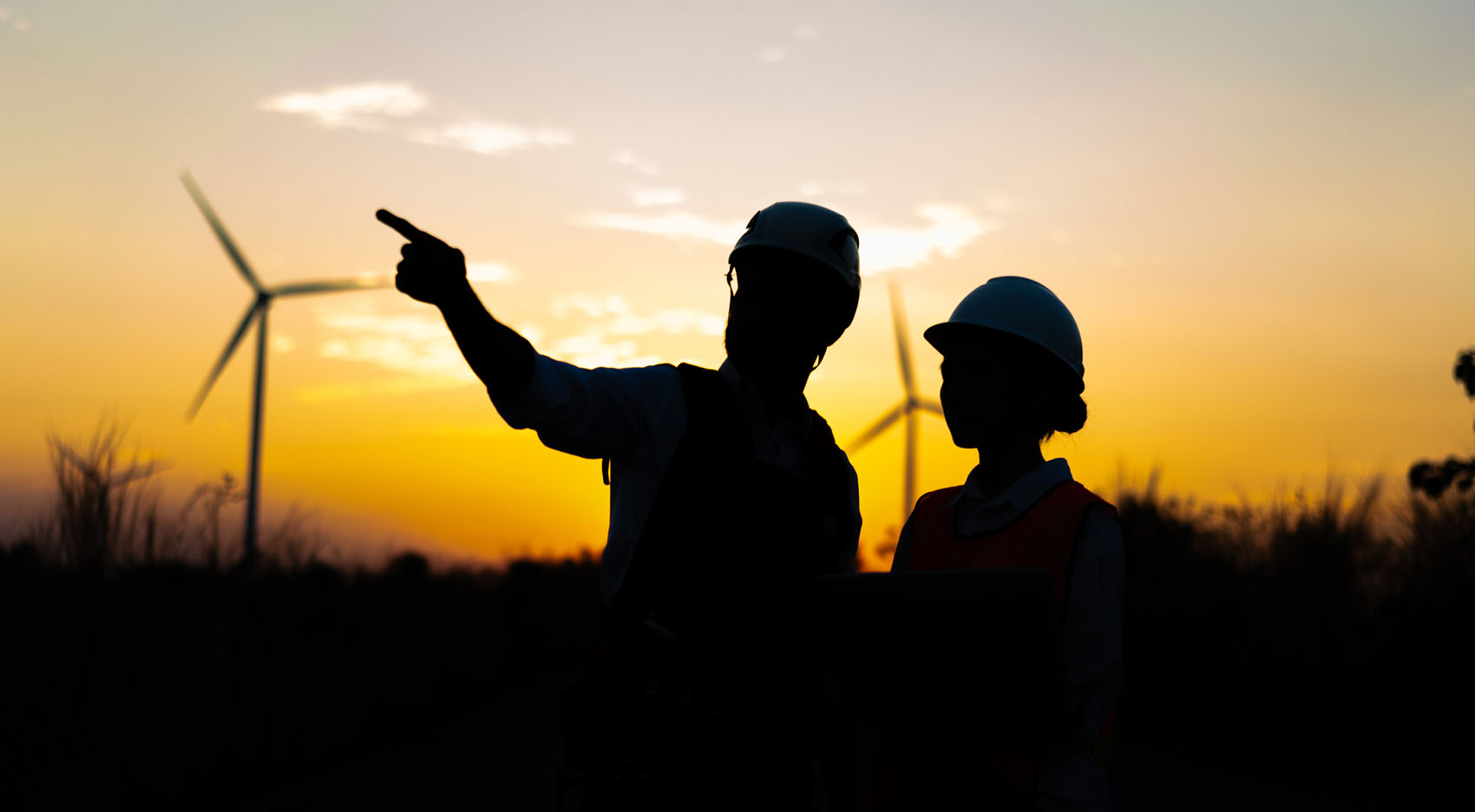 One person pointing in the distance as he talks to another person in a wind farm at sunset.
