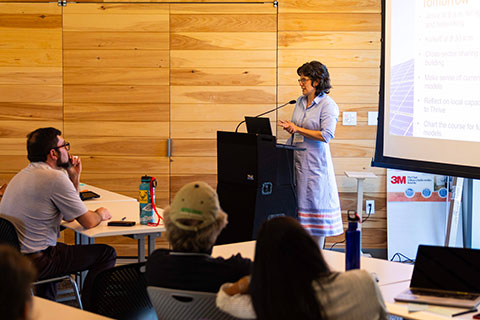 Suzanne standing at a podium with a screen behind her, speaking to an audience