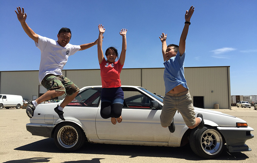 An adult and two children in a mid-air jump, with arms raised, in front of an old car.