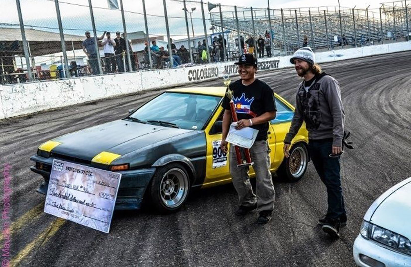 Two men stand beside a car with a ceremonial check leaning against the hood.