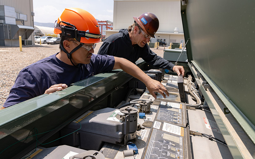Two people wearing hard hats work side by side on electrical switches.