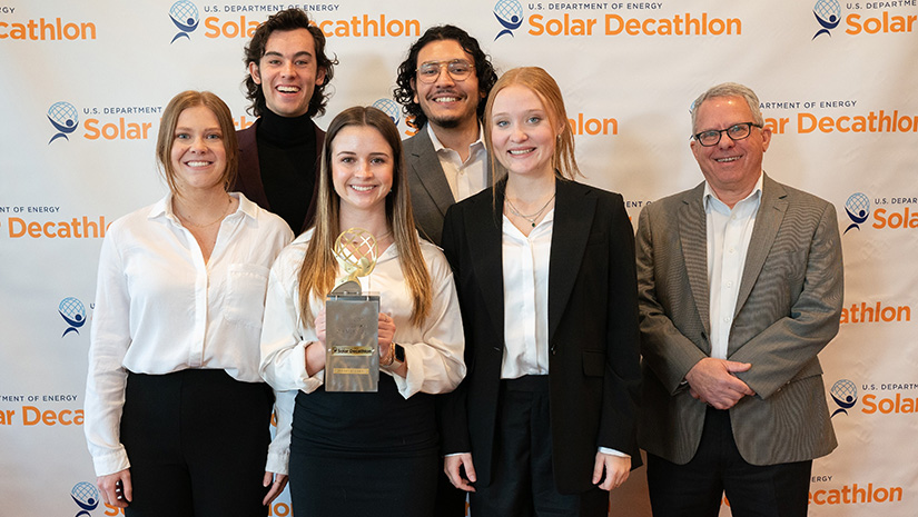 Students and a professor hold a trophy in front of a Solar Decathlon banner at an awards ceremony.