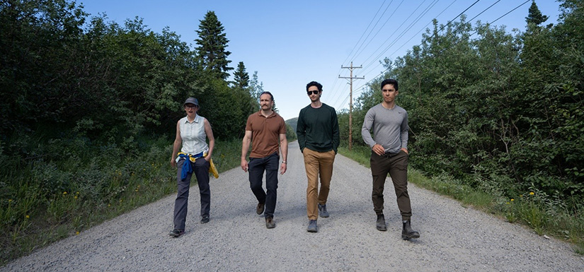 Four people walk down a gravel road surrounded by trees.