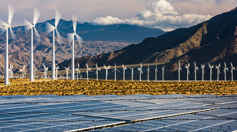 A solar energy farm in the forefront of a wind energy farm and mountains.