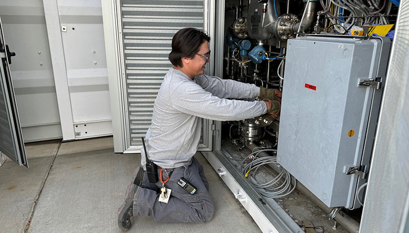 A researcher is kneeling on a concrete pad, wearing safety goggles and reaching into a side panel of large hydrogen research equipment.